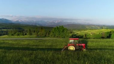 Traktör arka planda güzel yüksek dağlarla çimleri biçiyor, hava manzarası var. Dağ köyünde çiftçilik. Hasat sırasında traktör. Ekolojik tarım. 