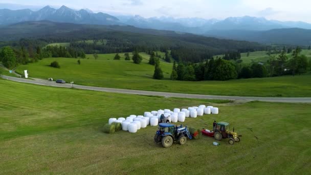 ヘイメイキングの空中ビュー 農業用バラーマシンとトラクター 山間の村で干し草の山とフィールド 背景の高い山の範囲 — ストック動画