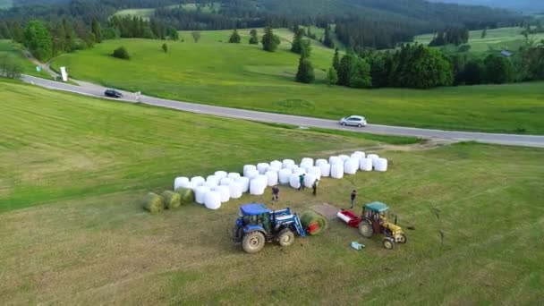 Letecký pohled na senoseč. Zemědělský balicí stroj a traktor. Farmáři pracující v úrodě. Pole plná balíků sena v horské vesnici. Krásný alpský pohled v pozadí. 