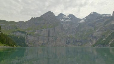 Dağlarda kar lekeleri ve hareket eden bulutlar olan bir gölün panoramik görüntüsü. Oeschinnensee. Kandertal. Bernese Oberland. Canton Bern. İsviçre. 