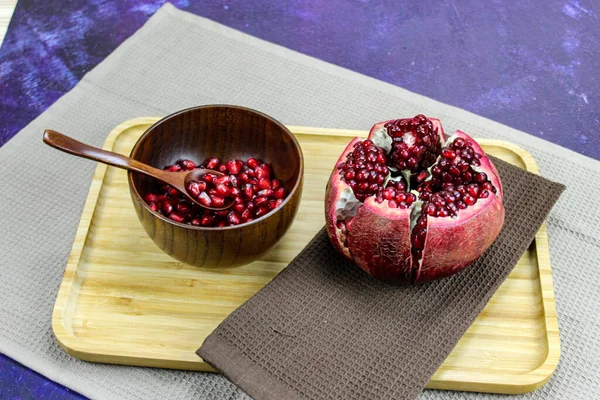 Wooden bowl containing wooden spoon and red pomegranate seeds. Pomegranate fruit open divided into five parts held together on a napkin / cloth on wooden tray on a blue background. Fruit pomegranate.