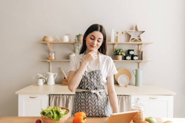 Önlüklü genç beyaz kadın modern mutfakta tablet bilgisayar kullanıyor, salata hazırlıyor, yemek tarifi okuyor...