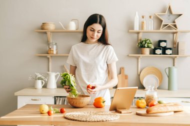Genç beyaz kadın modern mutfakta tablet bilgisayar kullanıyor, salata hazırlıyor, yemek tarifi okuyor..
