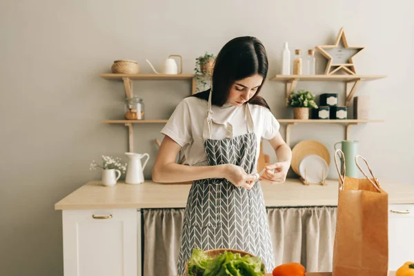 Young caucasian woman is tying an apron, preparation to cooking food, while standing in modern kitchen. Cooking food at home.