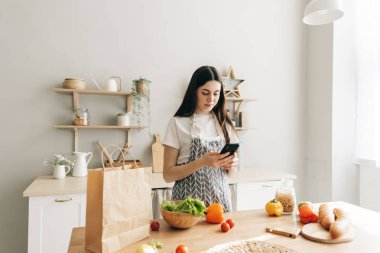 Önlüklü genç beyaz kadın modern mutfakta akıllı telefon kullanıyor, salata hazırlıyor, yemek tarifi okuyor..