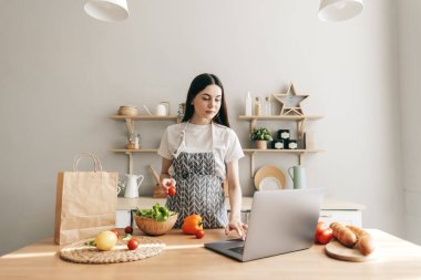 Önlüklü genç beyaz kadın modern mutfakta dizüstü bilgisayar kullanıyor, salata hazırlıyor, yemek tarifi okuyor..