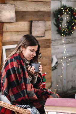 Ekoseli genç beyaz kadın verandada Noel ağacı ve süslemeleri olan sıcak bir içecek içiyor..