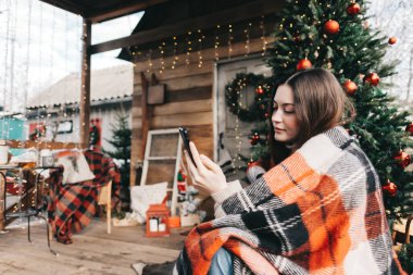 Arka bahçesinde Noel ağacı ve süslemeleriyle cep telefonu kullanan ekoseli beyaz bir kadın. Yüksek kalite fotoğraf