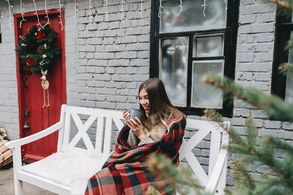 穿着格子衣服的年轻的高加索女人正坐在后院的长椅上喝着热饮 — 图库照片