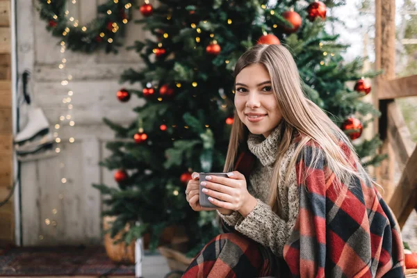 穿着格子花衣服的年轻的高加索女人正在阳台上喝着热饮料 上面有圣诞树和装饰品 — 图库照片