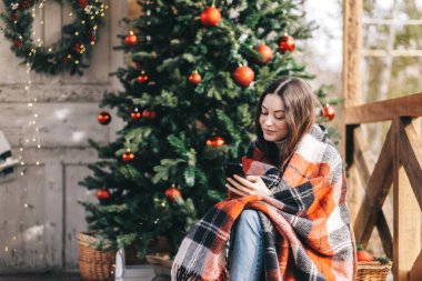 Arka bahçesinde Noel ağacı ve süslemeleriyle cep telefonu kullanan ekoseli beyaz bir kadın. Yüksek kalite fotoğraf