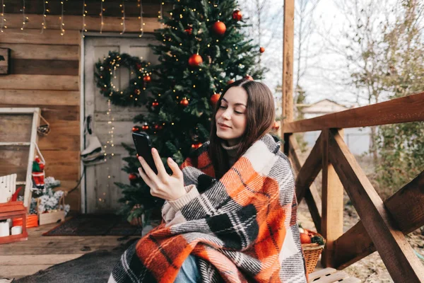 年轻的高加索女人穿着格子呢衣服 在后院用手机挂着圣诞树和装饰品 高质量的照片 — 图库照片