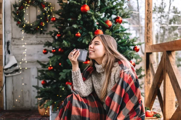 穿着格子花衣服的年轻的高加索女人正在阳台上喝着热饮料 上面有圣诞树和装饰品 — 图库照片