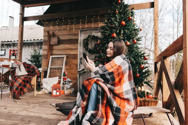 年轻的高加索女人穿着格子呢衣服 在后院用手机挂着圣诞树和装饰品 高质量的照片 — 图库照片