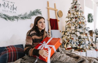 Mutlu beyaz kadın Noel ağacının yakınındaki bir yatakta otururken elinde büyük bir Noel hediyesi tutuyor..