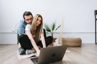 Genç çift dizüstü bilgisayar kullanıyor, bilgileri tarıyor ve modern oturma odasındaki dizüstü bilgisayarlara bakıyor....