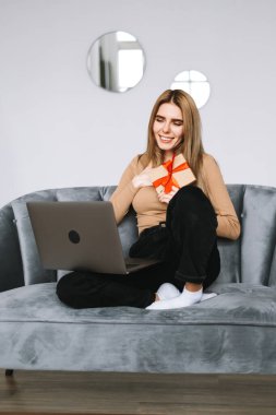 Mutlu genç bir kadın video görüşmesi ya da sohbet sırasında dizüstü bilgisayarın önünde hediyesini açıyor. İnternette kutluyor. Mesafe ilişkileri, kutlamalar ve yaşam tarzı kavramı.