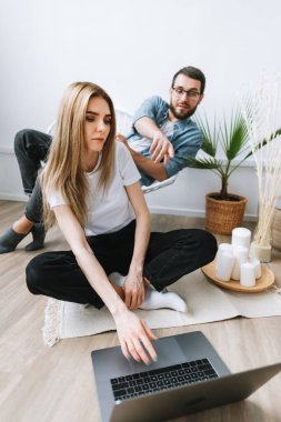 Genç çift dizüstü bilgisayar kullanıyor, bilgileri tarıyor ve modern oturma odasındaki dizüstü bilgisayarlara bakıyor..