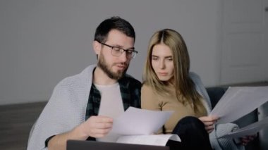 Genç çift dizüstü bilgisayarlarıyla finansman yönetiyor. Oturma odasındaki banka hesaplarına bakıyorlar. Kadın ve erkek birlikte belgelere bakar. Bütçe harcamaları planlanıyor.