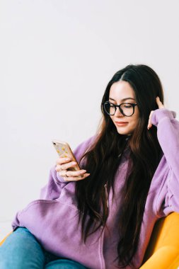 Genç beyaz kadın cep telefonu kullanıyor, oturma odasında koltukta oturuyor..