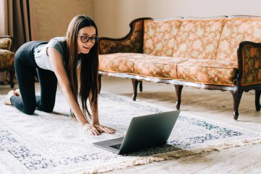Güzel esmer fitness kadını dizüstü bilgisayarında esneme egzersizleri yapıyor, evde yoga yapıyor. Formda ve sağlıklı kalmak.