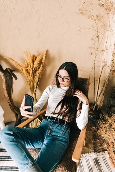 Young caucasian woman holding smartphone looking on cell using mobile phone technology at home, taking selfie for social media or video calling in app virtual chat sitting on armchair.