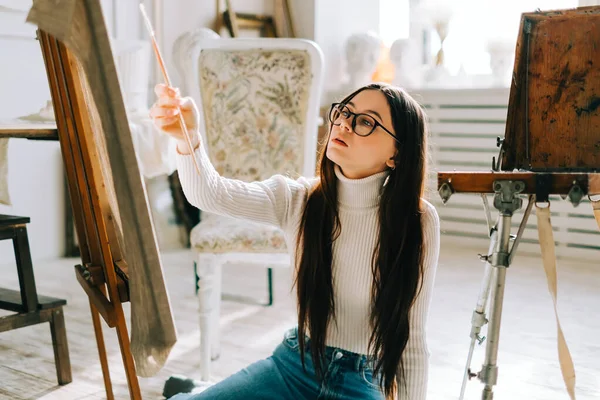 若い白人女性アーティストが床に座り 絵筆を持ってキャンバスに絵を描き 自宅のスタジオで仕事をしています — ストック写真