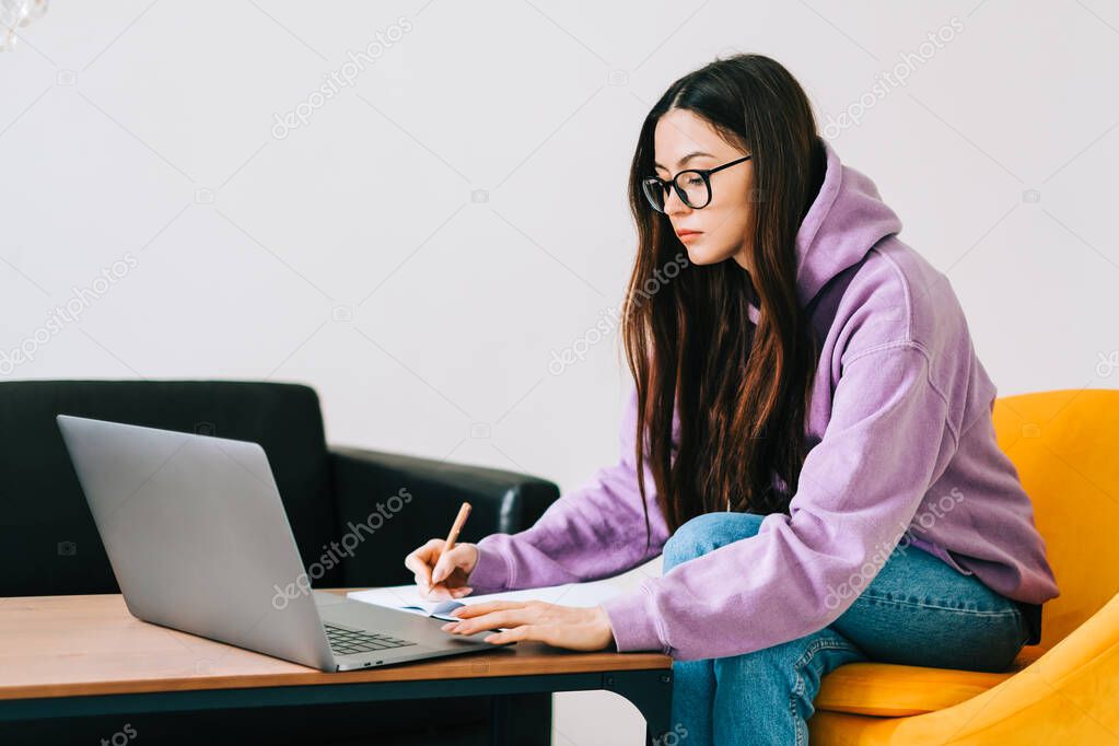 Focused caucasian young woman college student in eyeglasses studying with laptop, distantly preparing for test exam, writing essay doing homework at home, distantly education concept.