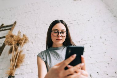 Beyaz tuğlalı duvarın yanında cep telefonu kullanan gözlüklü genç beyaz kadın..