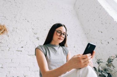 Beyaz tuğlalı duvarın yanında cep telefonu kullanan gözlüklü genç beyaz kadın..