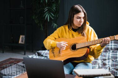 Beyaz bir kadın evde gitar çalmayı öğreniyor, dizüstü bilgisayar kullanarak online kurslar izliyor, yatakta oturuyor. Uzaklık eğitimi kavramı