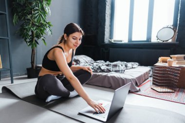 Güzel esmer fitness kadını dizüstü bilgisayarında esneme egzersizleri yapıyor, evde yoga yapıyor. Formda ve sağlıklı kalmak.