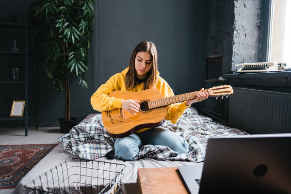 Young caucasian woman learning to play guitar at home, watching online courses using a laptop, sitting on the bed. Distance education concept