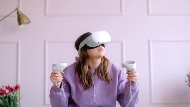 Tânăra femeie caucaziană care stă pe pat și folosește căștile VR, deține controlere și caută în realitatea virtuală . — Videoclip de stoc