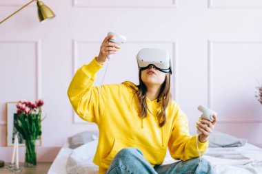 Genç beyaz kadın yatakta oturuyor ve VR kulaklık takıyor. Kontrolörleri tutuyor ve sanal gerçekliğe bakıyor..