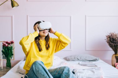 Kafkasyalı genç bir kadın VR kulaklık takıyor, gözlüklere dokunuyor ve sanal gerçekliğe bakıyor, evdeki yatakta oturuyor..