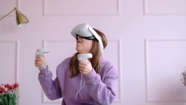 Jonge blanke vrouw die op het bed zit en VR-headset gebruikt, controllers vasthoudt en in virtual reality kijkt. — Stockvideo