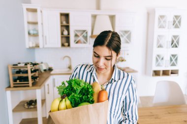 Genç gülümseyen beyaz kadın modern mutfakta taze sebze ve baget dolu eko alışveriş çantası tutuyor..
