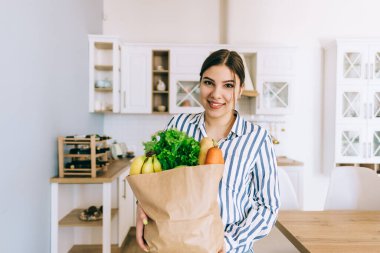 Genç gülümseyen beyaz kadın modern mutfakta taze sebze ve baget dolu eko alışveriş çantası tutuyor..