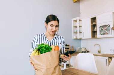 Genç gülümseyen beyaz kadın modern mutfakta akıllı telefon kullanıyor. Masada taze sebzeler var. İnternetten gıda ve market alışverişi.