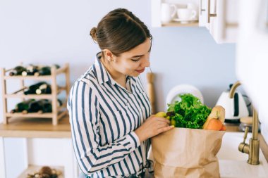 Genç beyaz kadın mutfaktaki kese kağıdından sebze alıyor..