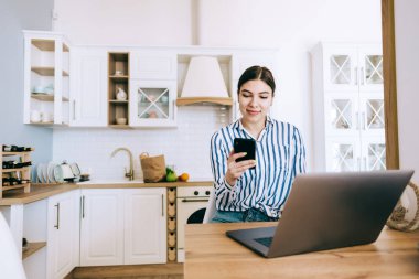 Evde çalışan, evdeki mutfakta oturan, cep telefonu ve dizüstü bilgisayar kullanan beyaz bir kadın..