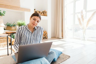 Genç beyaz kadın rahat oturma odasında dizüstü bilgisayarda çalışıyor, yerde yastıklarla oturuyor..