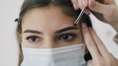 Usta, kaş şeklini bir güzellik salonunda maske takan genç bir kadına boyuyor. Yakın plan.