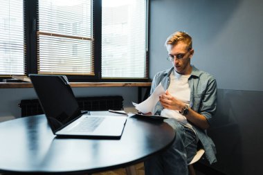 Pensive caucasian man working with laptop at home office and looking on paper documents, calculating bills.	