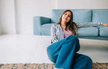 Genç, sakin, Kafkasyalı kadın dinleniyor, modern oturma odasındaki kanepenin yanında oturuyor, evde dinlenen tembel, mutlu kadın.