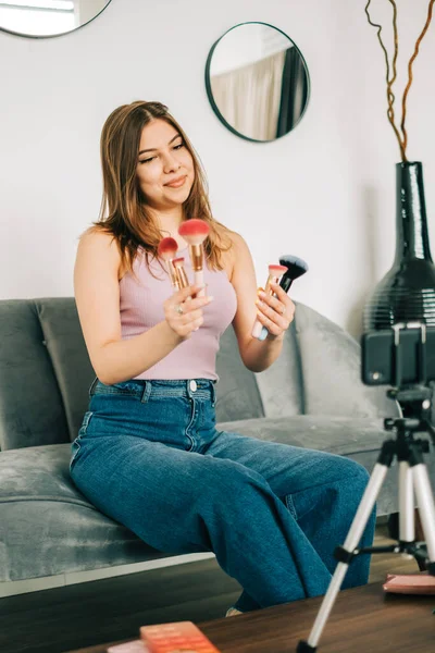 迷人的高加索年轻女性视频博主通过她的博客在社交媒体上展示美容产品 手持化妆刷和阴影看着安装在三脚架上的智能手机相机 — 图库照片