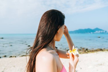 Genç beyaz kadın güneşli bir günde kumlu bir sahilde mavi suyla yürüyor..