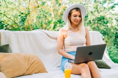 Dışarıda dizüstü bilgisayar kullanan beyaz bir kadın yaz günü arka bahçede bir bankta oturuyor..