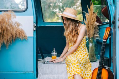 Genç, beyaz bir kadın dışarıda bir minibüste otururken dizüstü bilgisayar kullanıyor..